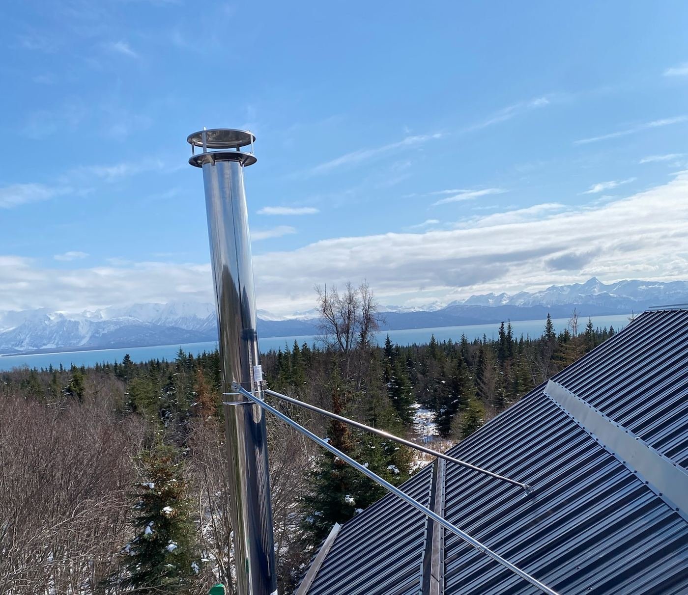 Stainless chimney installation on a metal roof overlooking Kachemak Bay and snow-capped mountains