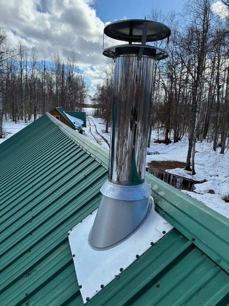 Stainless chimney on a green metal roof surrounded by snowy birch forest